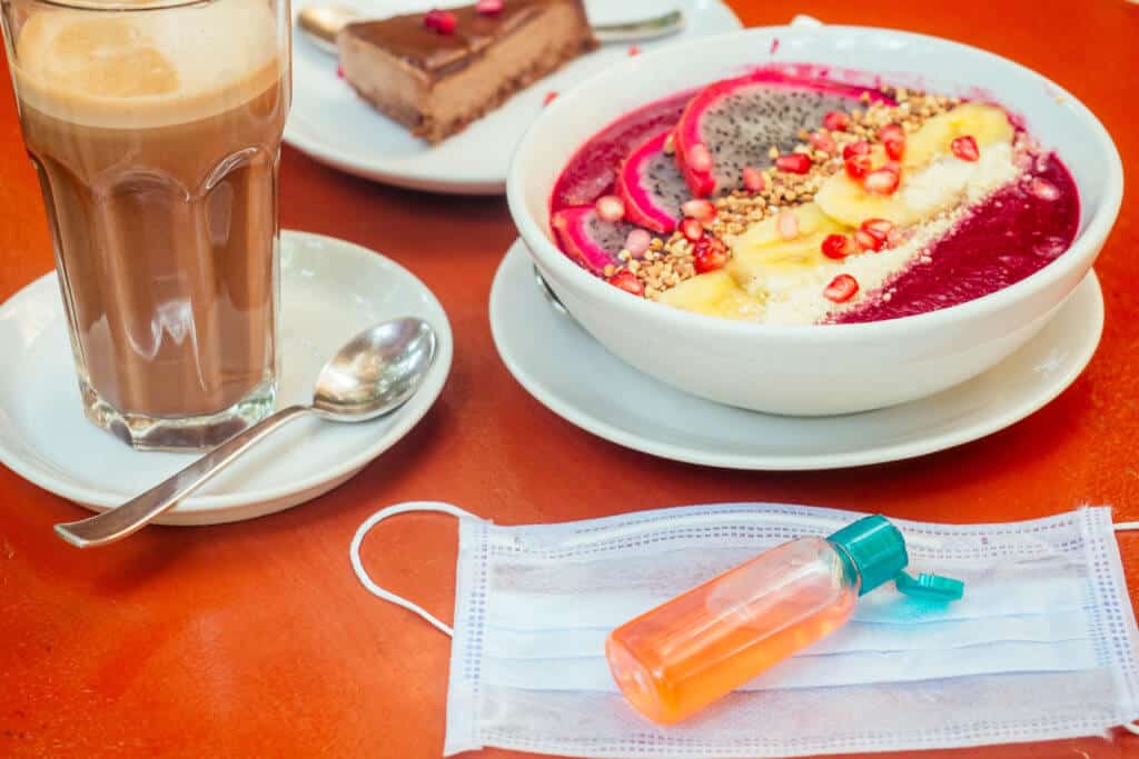 woman using antibacterial gel on hands before start to eating her healthy food in tropical asian cafe . smoothie bowl , raw cake and coffee on table with sanitizer on bottle